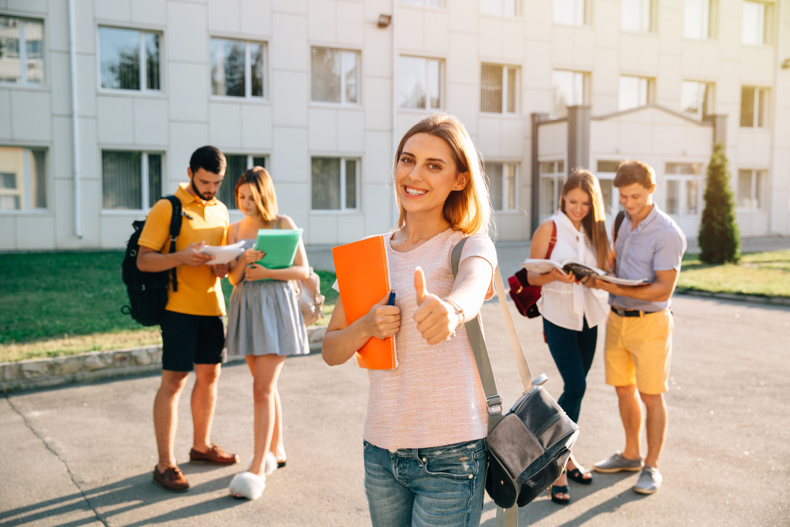 Happy beautiful girl standing with note books and backpack showing thumb up and smiling, standing near university building, her friends are behind Happy beautiful girl standing with note books and backpack showing thumb up and smiling, standing near university building, her friends are behind